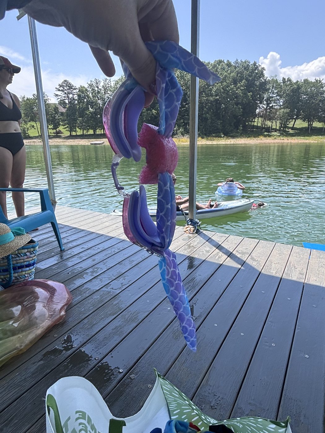 Mermaid-print swim goggles with a pink Goggle Bean held up over a sunny lake dock