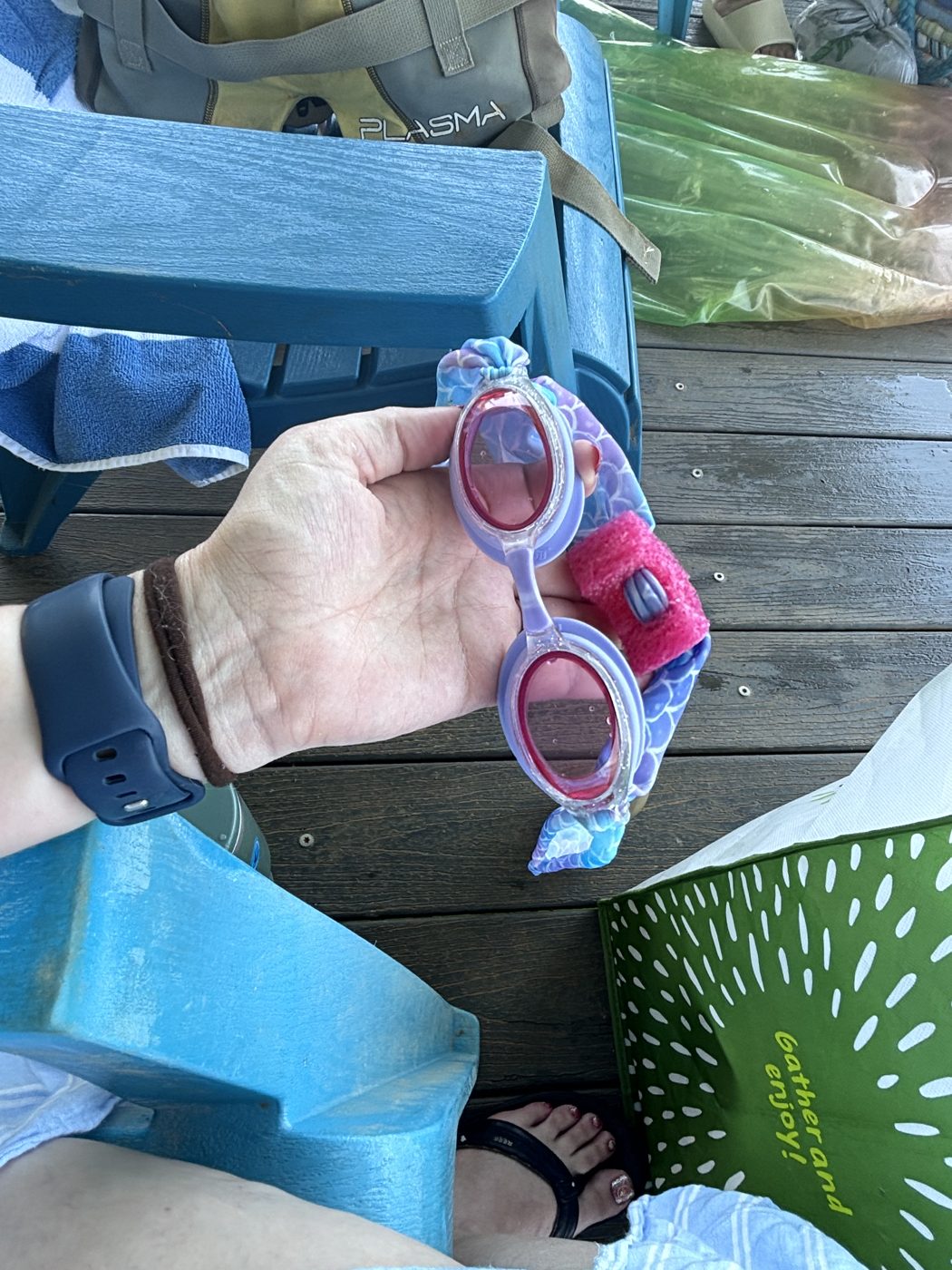 A hand holding swim goggles fitted with a pink Goggle Bean on a lake dock